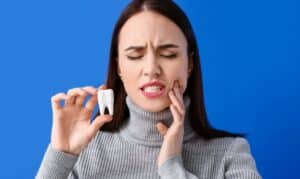 Woman experiencing tooth pain, holding a knocked-out tooth against blue background.